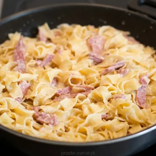 Kremowy makaron tagliatelle w sosie śmietanowym z paskami podsmażonej szynki, posypany żółtym serem.