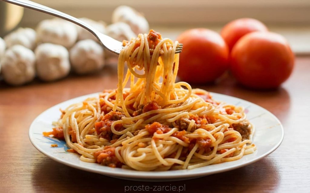 Duży talerz spaghetti po polsku z gęstym sosem mięsnym, tartą marchewką i posypką z ziół.