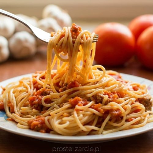 Duży talerz spaghetti po polsku z gęstym sosem mięsnym, tartą marchewką i posypką z ziół.