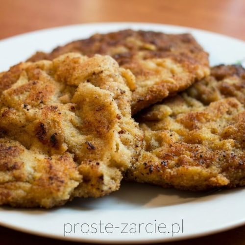 Złocisty, chrupiący kotlet schabowy podany z ziemniakami i koperkiem na białym talerzu.
