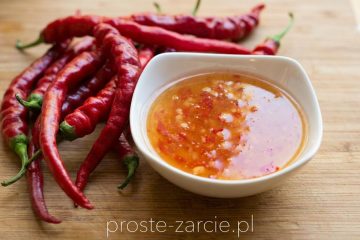 Szklisty, gęsty domowy słodki sos chili z widocznymi kawałkami papryczki w małej miseczce, obok leży świeże chili.