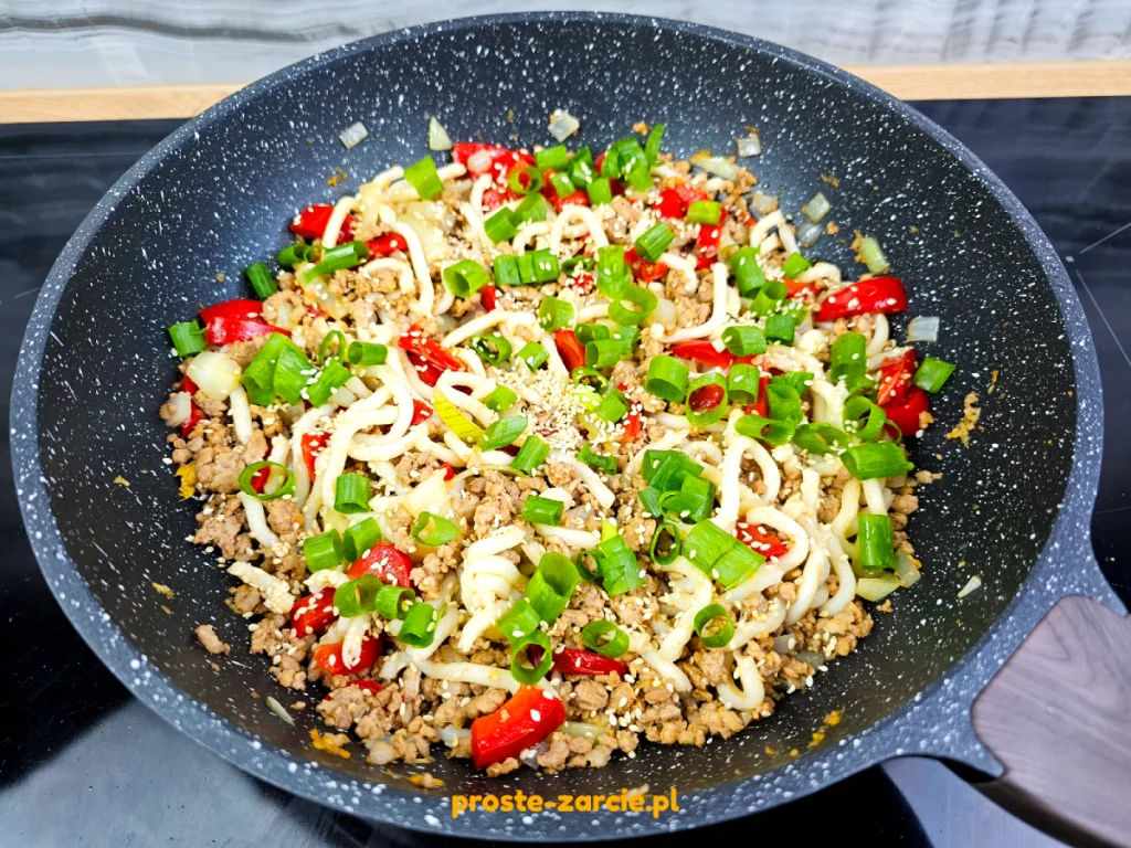 Miska pełna grubego makaronu udon z mieloną wieprzowiną, papryką, posypana szczypiorkiem i uprażonym sezamem.