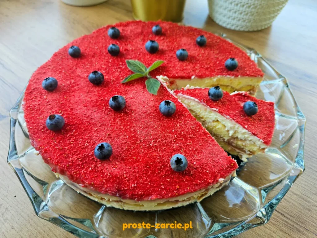Kawałek tiramisu truskawkowego podany na talerzu, polany sosem owocowym i udekorowany mięta oraz borówkami.
