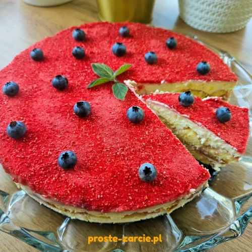 Kawałek tiramisu truskawkowego podany na talerzu, polany sosem owocowym i udekorowany mięta oraz borówkami.
